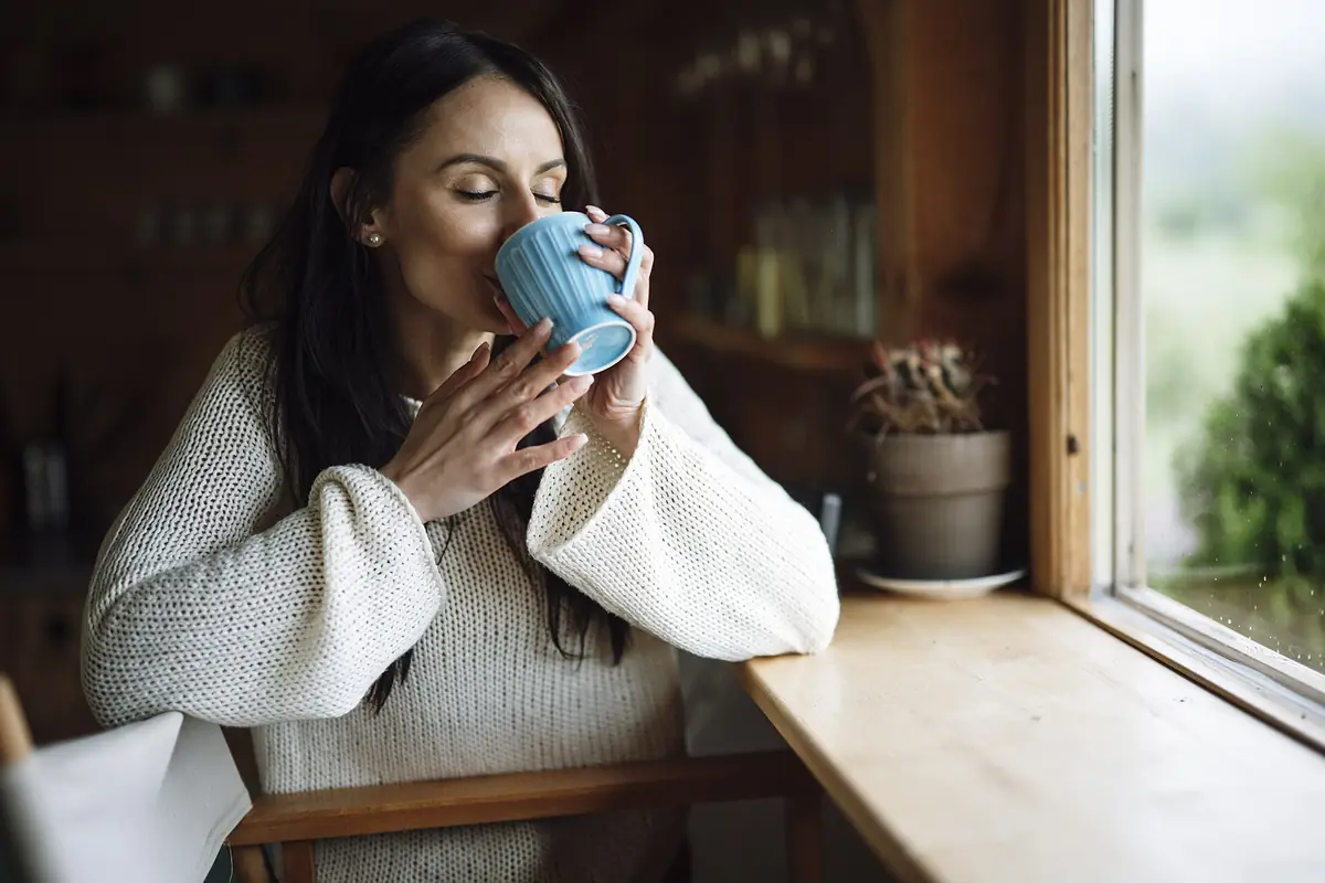A woman sipping from a blue cup while sitting by a window in a cosy room