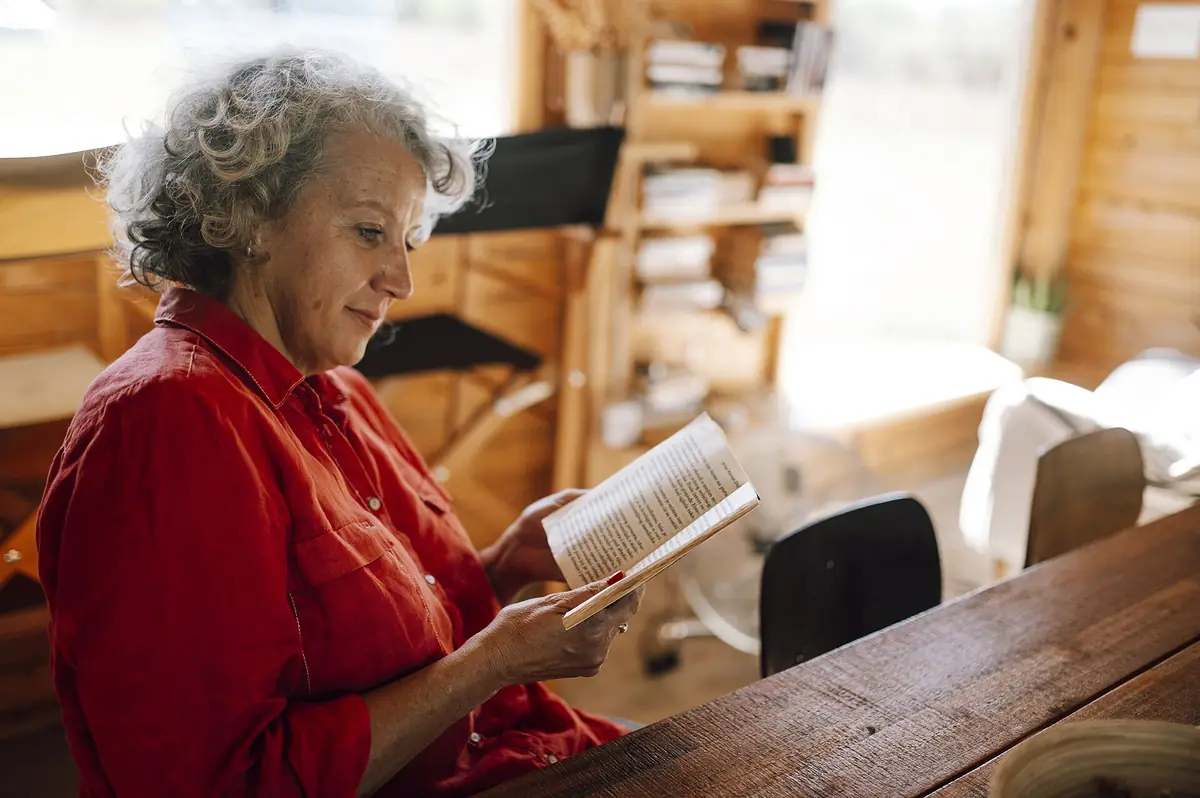 An older woman reading a book at a wooden table in a cosy home