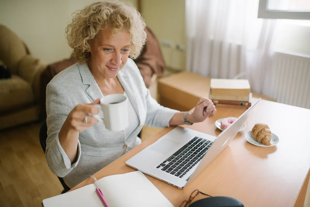 A woman in her sixties with a cup of coffee working on her laptop at home